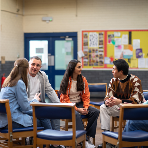 A diverse group of adults sits in a circle having a supportive discussion in a community center or group therapy setting. They appear engaged and comfortable, suggesting a group counseling or mental health session.